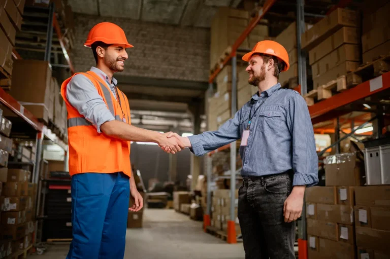 Dois homens em ambiente logístico se cumprimentam, simbolizando o acordo de um contrato de locação de galpão.
